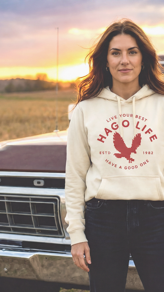 A woman stands outdoors in front of a vintage pickup truck at sunset, wearing a cream hoodie with a red graphic that reads 'HARPOULIFE' and black pants.
